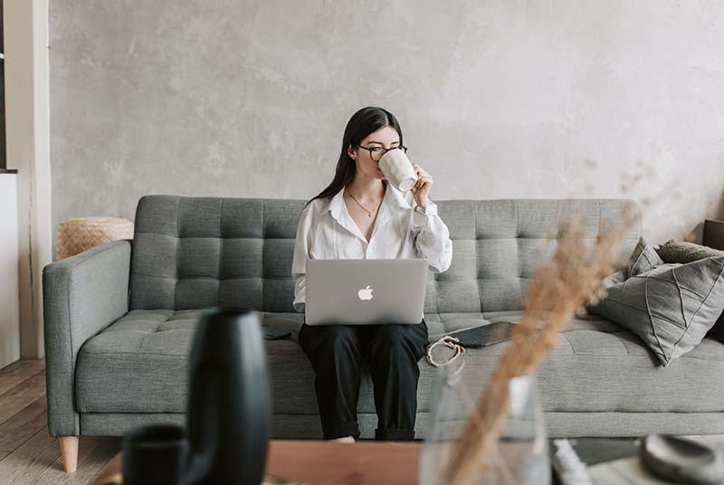 A woman wearing black glasses is sitting on a gray couch, looking down at her silver Apple laptop on her lap, and drinking from a mug of coffee from her left hand.