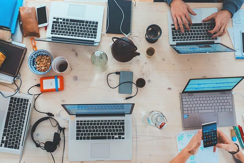 A group of people have their laptops on a desk, with clutter of headphones, snacks, and notebooks.