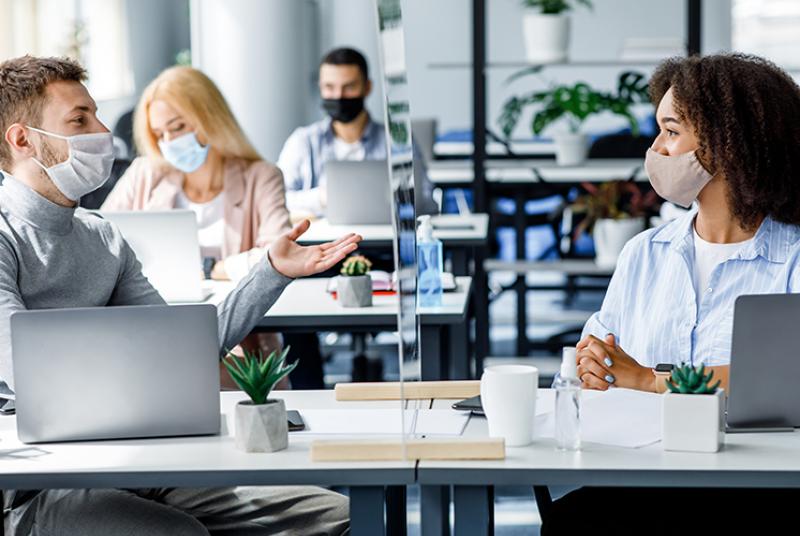 In an office setting, people are working at their laptops, wearing face masks and at separated desks. At the front of the picture, a white man is wearing a face mask and talking to his coworker, a black woman wearing a face mask. Their desks are separated