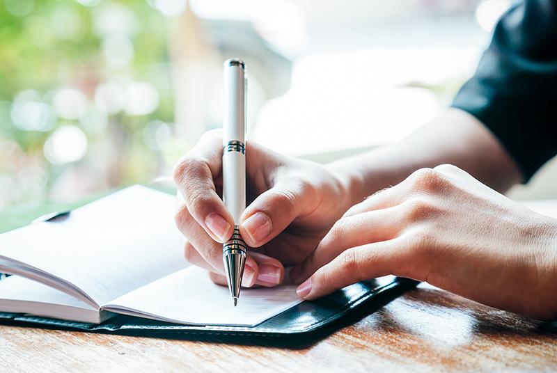 Hands writing in a notebook with green dappled scenery in the background